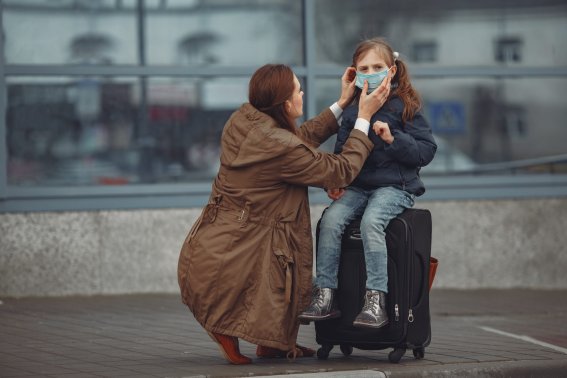 A European mother in a respirator with her daughter are standing near a building.The parent is teaching her child how to wear protective mask to save herself from virus.