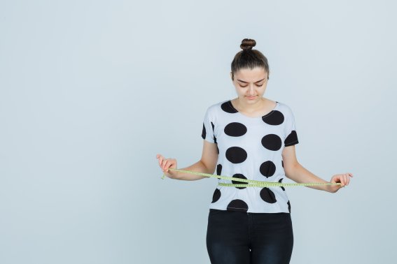 young woman holding tape measure, measuring waist size in white t-shirt and black pants and looking focused. front view.