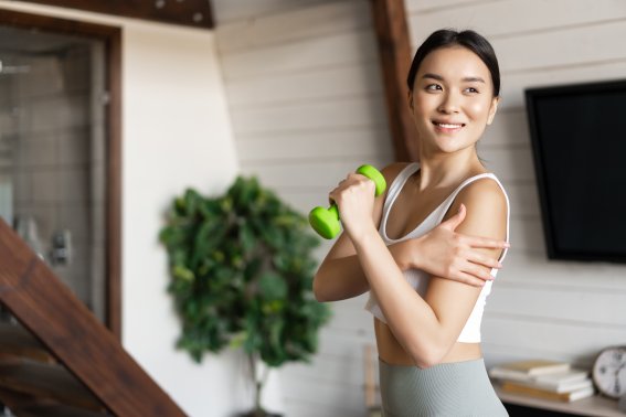 Active and healthy asian girl with fit body doing fitness exercises at home, lifting dumbbells and touching her biceps, workout in living room.